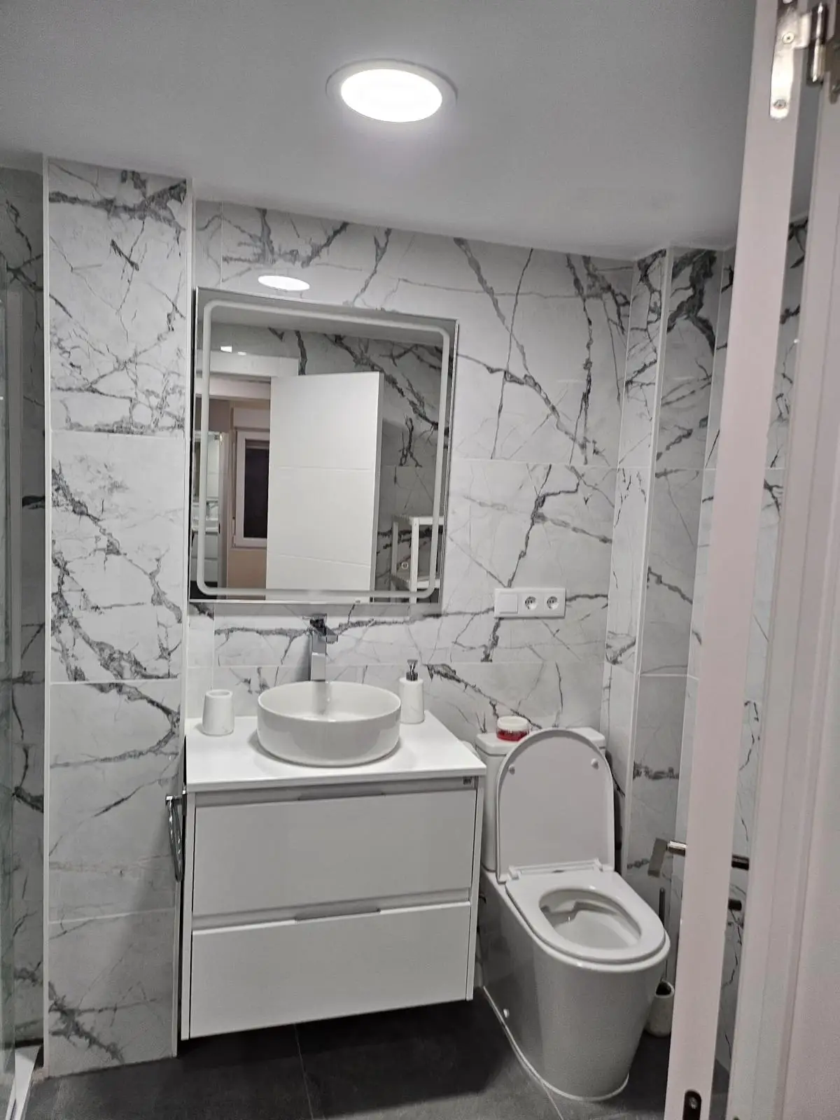 Reforma de baño con revestimiento cerámico efecto mármol, lavabo sobre encimera y espejo retroiluminado en Madrid